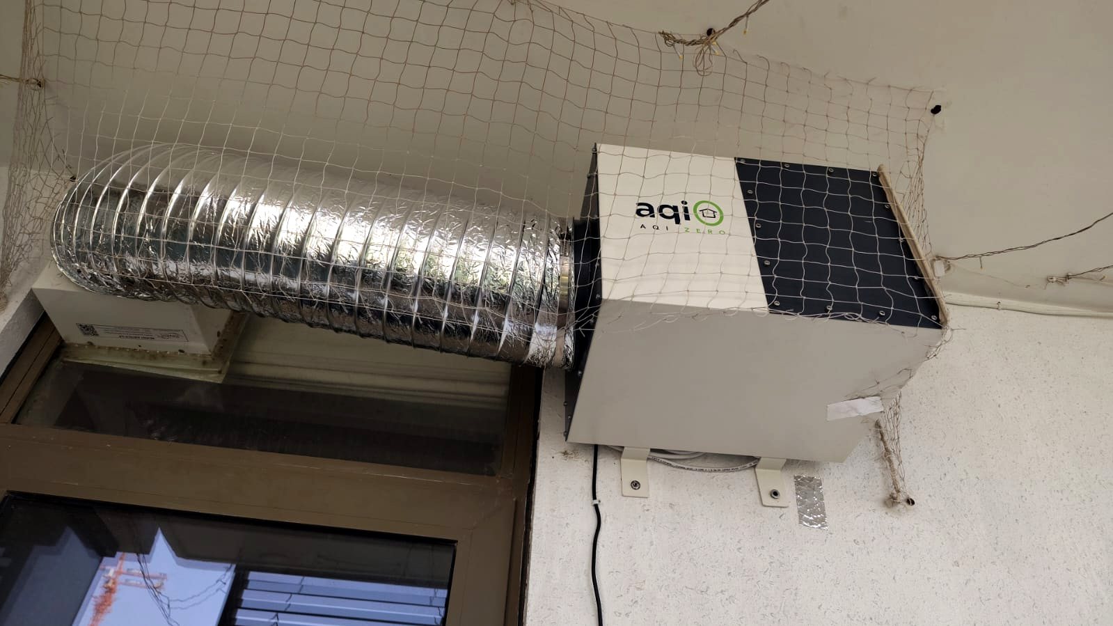aqi0 wall-mounted unit behind balcony netting with a duct above a window