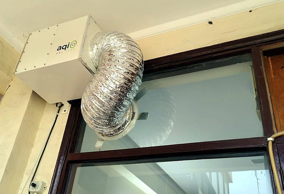 An aqi0 unit mounted high on a wall, its silver duct passing through a glass pane