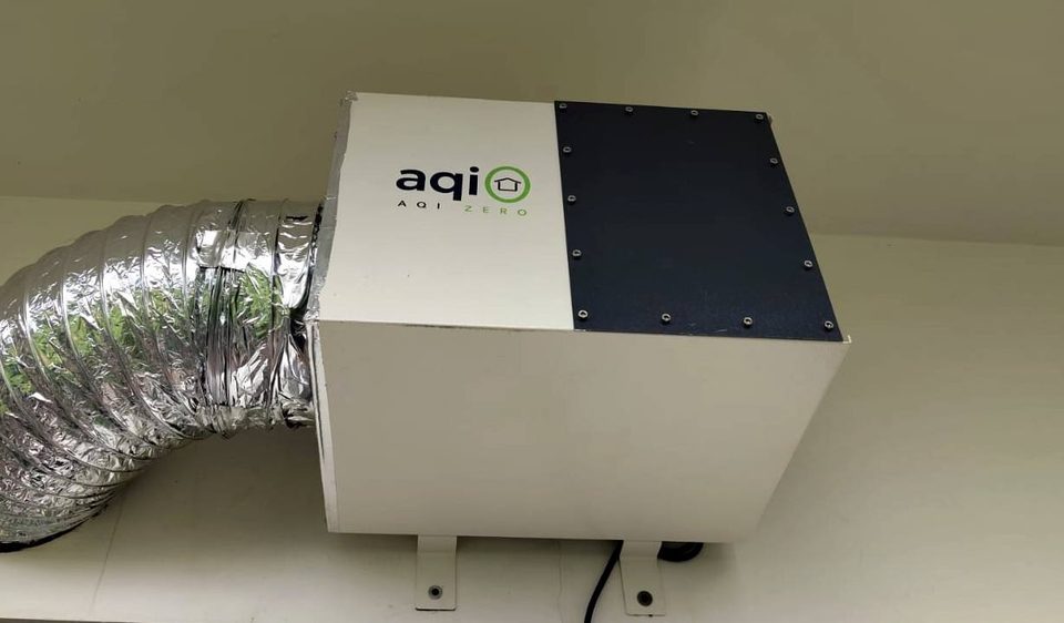 Close-up of an aqi0 unit: white sheet-metal body, black front plate fixed with screws, silver duct on the left