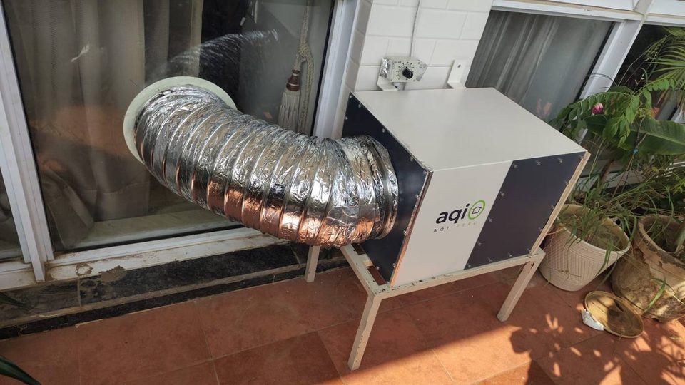 An aqi0 unit on a metal floor stand on a balcony, its silver duct curving into a round opening in the glass door
