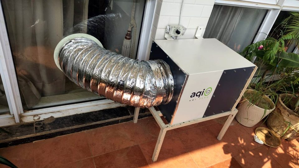 An aqi0 unit on a metal floor stand on a balcony with a terracotta floor, its silver duct running to the glass door beside it