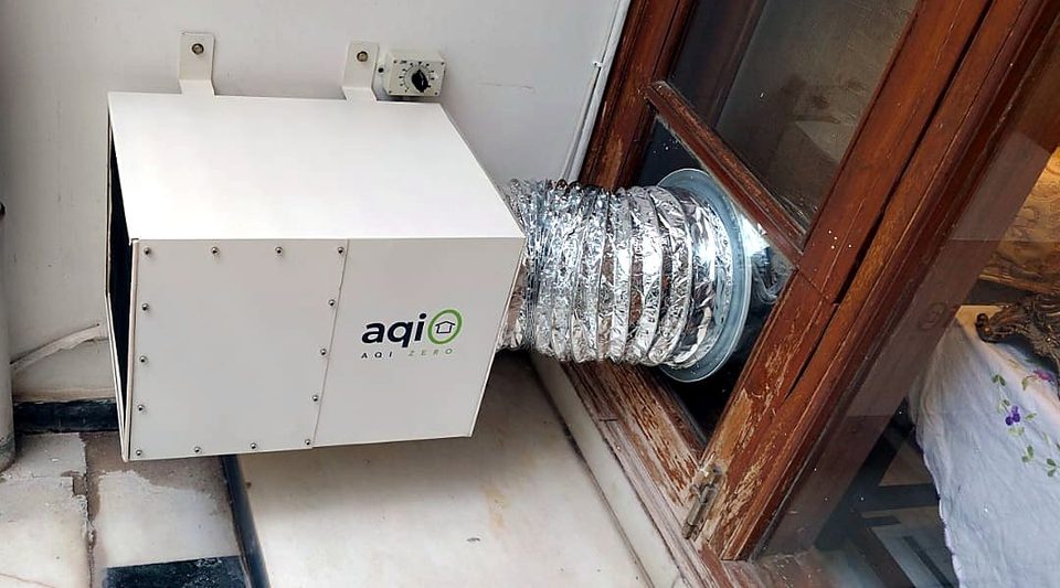 An aqi0 unit wall-mounted beside a wooden window, its flexible duct running into a panel fitted in the window frame